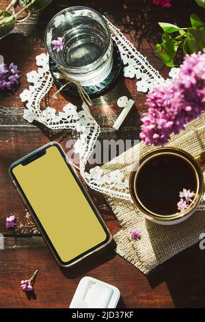 Stile di vita estetico piatto, cuffie senza fili, mockup schermo del telefono, decorazioni fiori lilla, tazza di caffè e bicchiere d'acqua. Ascoltare musica o audiolibro, surf stile di vita telefono Foto Stock