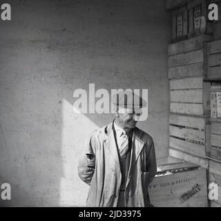 1940s, storico, un lavoratore di negozio/magazzino maschile sorridente, indossando una camicia e una cravatta sotto un'opera o un cappotto di tatta, pieno di fori, e un cappuccio di stoffa, in piedi per una foto all'interno, accanto ad una pila di casse di legno, Bramsall, Stockport, Manchester, Inghilterra, REGNO UNITO. Foto Stock