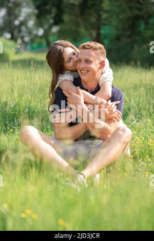 Coppia giovane carina seduta sull'erba, baciare. Giovane uomo e donna che si godono giorno d'estate. Gente felice nell'amore, luna di miele Foto Stock