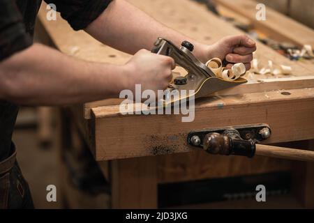 Primo piano le mani del falegname che lavorano con legno, tagliando, affilando in aereo, trucioli che volano intorno al banco di lavoro. Affari Foto Stock