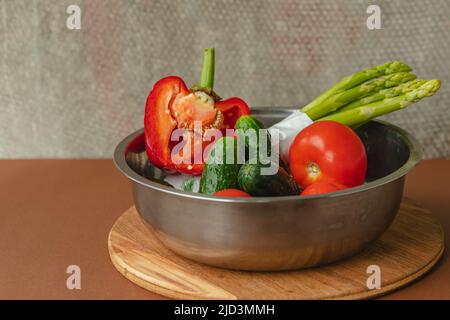 Le verdure si trovano in una ciotola di metallo pomodori, asparagi, cetrioli, peperoni rossi . su un asse di legno e sfondo marrone. Sfondo grigio posteriore. Luogo Foto Stock