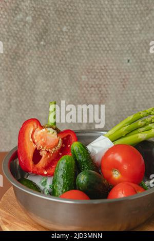 Le verdure si trovano in una ciotola di metallo pomodori, asparagi, cetrioli, peperoni rossi . su un asse di legno e sfondo marrone. Sfondo grigio posteriore. Luogo Foto Stock