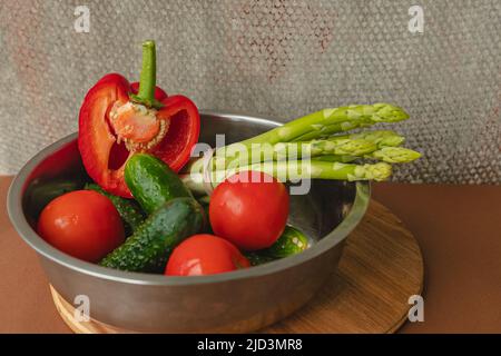 Le verdure si trovano in una ciotola di metallo: Pomodori, asparagi, cetrioli, peperoni rossi . su un asse di legno e sfondo marrone. Sfondo grigio posteriore. Luogo Foto Stock