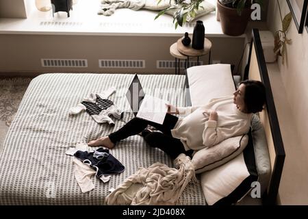 Una donna incinta si trova sul letto di fronte a un computer portatile e guarda un bel film. Il concetto di un momento piacevole a casa. Messa a fuoco soft. Vista dall'alto. Foto Stock