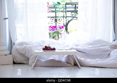 Un piccolo tavolo con fragole e un cavalletto di vetro vicino al letto sul pavimento vicino al balcone aperto nell'appartamento, camera da letto interna, colazione in letto Foto Stock