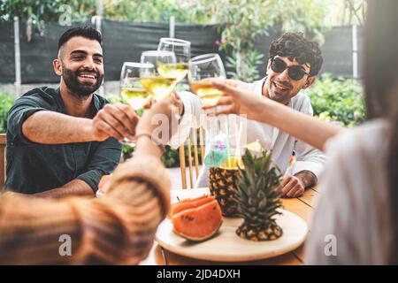 Un gruppo giovane di amici che ha un brindisi celebrativo che si abbinano a bicchieri bianchi e che mangia frutta fresca e succosa seduti in una terrazza di un caffè o di un ristorante Foto Stock