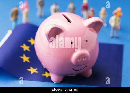 Piggy Bank, bandiera dell'UE e giocattoli di plastica su sfondo colorato, il concetto di reddito nell'Unione europea Foto Stock