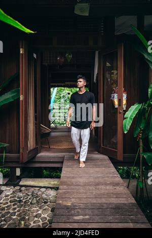 giovane uomo che cammina su un sentiero di legno dal piccolo ingresso tropicale casa a bali indonesia Foto Stock