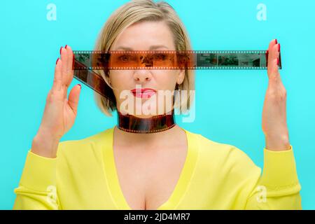 Una donna bionda alla moda tiene in mano un film fotografico, un film negativo su sfondo turchese. Vivace ritratto femminile alla moda Foto Stock