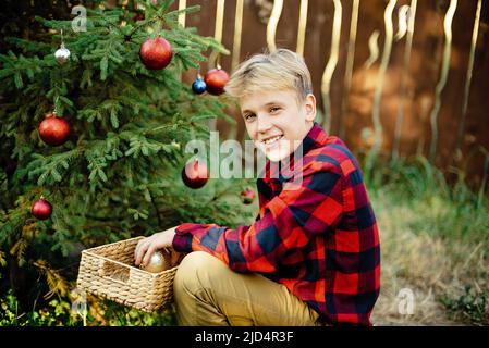 Natale nel mese di luglio. Bambino in attesa di Natale in legno in estate. Ritratto di ragazzo in abito rosso decorando albero di natale. Vacanze invernali e concetto di gente. Buon Natale e buone feste Foto Stock