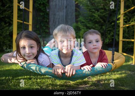 tre bambini, una ragazza e ragazzi, fratelli e sorelle, oscillano stesi su un altalena nel parco giochi. Infanzia allegra, incuria, vacanze estive, gioia Foto Stock