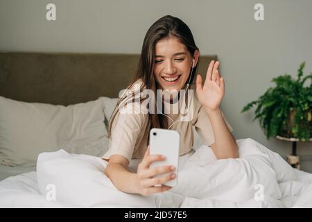 Donna seduta a letto dopo essersi svegliata e aver parlato con una videochiamata utilizzando il telefono cellulare e le cuffie Foto Stock