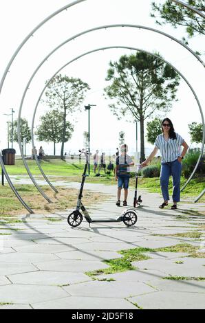 Felice stile di vita familiare e concetto di vacanza. Madre, ragazzino che guida scooter, a piedi nella città vecchia, strada. Risate in una giornata estiva di sole. Divertimento Foto Stock