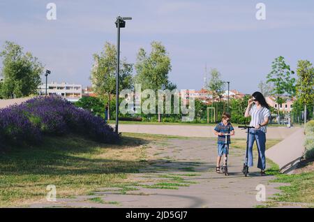 Felice stile di vita familiare e concetto di vacanza. Madre, ragazzino che guida scooter, a piedi nella città vecchia, strada. Risate in una giornata estiva di sole. Divertimento Foto Stock