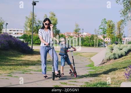 Felice stile di vita familiare e concetto di vacanza. Madre, ragazzino che guida scooter, a piedi nella città vecchia, strada. Risate in una giornata estiva di sole. Divertimento Foto Stock