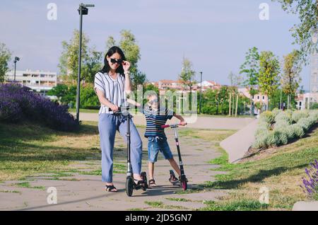 Felice stile di vita familiare e concetto di vacanza. Madre, ragazzino che guida scooter, a piedi nella città vecchia, strada. Risate in una giornata estiva di sole. Divertimento Foto Stock