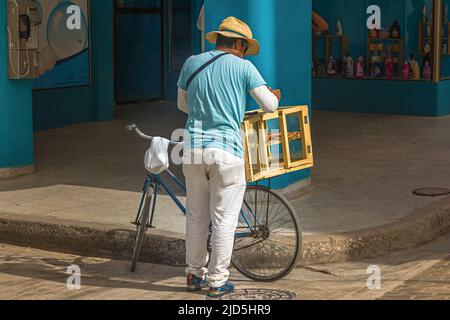 venditore di strada con un cappello di paglia, pantaloni bianchi e T-shirt blu chiaro e una bicicletta vende prodotti da forno nella città di Camagüey, Cuba Foto Stock