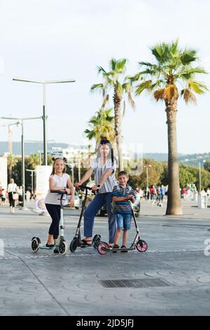 Felice stile di vita familiare e concetto di vacanza. Madre, ragazzino, ragazza , scooters a cavallo, camminando nella città vecchia, strada. Risate in una giornata estiva di sole. Foto Stock