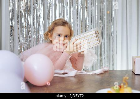 Vacanze per bambini. Felice, ragazza festivamente vestita con stelle scintillanti sulle sue guance sta tenendo la scatola del regalo nelle sue mani con anticipazione, palloncini sono Foto Stock