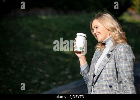 Bionda affascinante con lunghi capelli ricci in giacca in tweed grigio-blu e maglione in cashmere abbinato, tenendo una tazza di tè nelle sue mani mentre cammina al sole Foto Stock