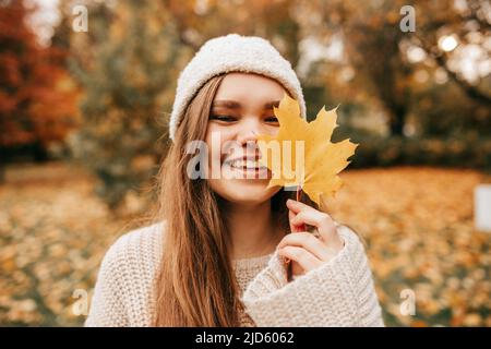 Ragazza sorridente carina in cappello a maglia, maglione nella foresta autunnale, tenendo in mano la foglia di acero, camminando nel parco cadente Foto Stock