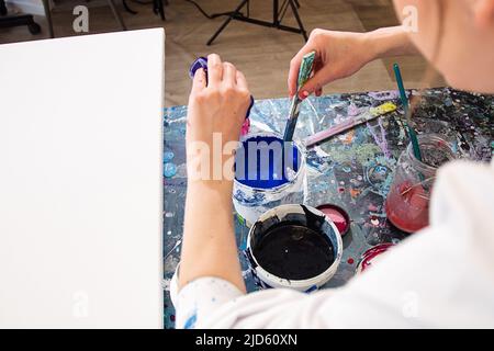 Foto ritagliata di mani di donna in accappatoio bianco versando vernice da piccola bottiglia di plastica in secchio con blu, mescolando. Foto Stock