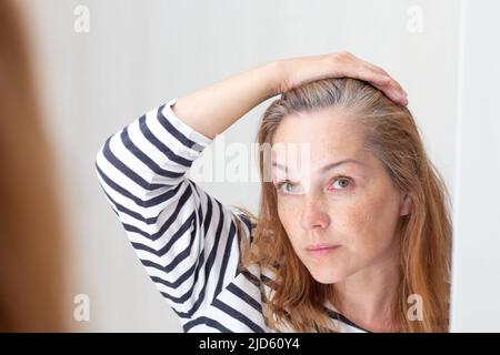 Donna di mezza età che guarda i capelli grigi in riflessione specchio su crescere fuori radice Foto Stock
