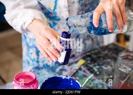 Foto ritagliata di mani di donna in accappatoio coperto con macchie versando acqua dal vaso di vetro in una piccola bottiglia di plastica. Foto Stock
