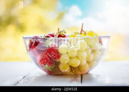 Recipiente in vetro trasparente riempito con fragole e uva appena raccolte, lavato in acqua. Cielo luminoso e poco chiaro e gli alberi sullo sfondo. Orga estiva Foto Stock