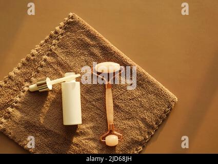 Set per la cura del viso a casa su un asciugamano marrone. Rullo per massaggio viso al quarzo rosa o gua sha e siero in una bottiglia di vetro bianco e pipetta per la cura del viso. Foto Stock