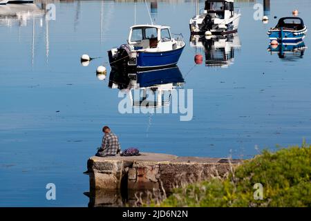 Riflessione silenziosa. Un uomo seduto sul porto in un'atmosfera riflettente, con barche che proiettano riflessi sul mare a specchio in una luminosa mattinata di sole Foto Stock