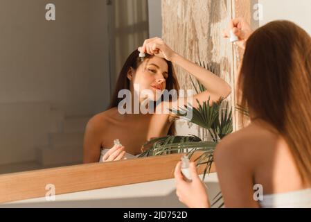 Una bella giovane donna usa un siero idratante anti-invecchiamento del viso in bagno. Cura della pelle del corpo e concetto di applicazione cosmetica Foto Stock