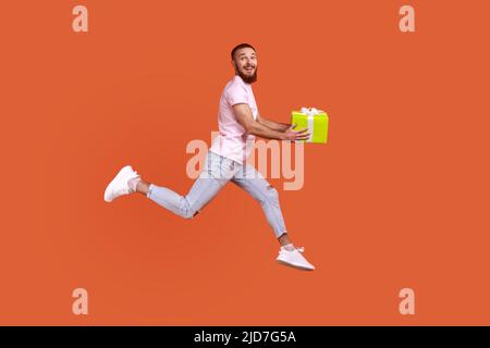 Ritratto a tutta lunghezza di uomo bearded ispirato che saltano in aria o che corre rapidamente veloce per dare il presente box, esprimendo la felicità, indossando la T-shirt rosa. Studio interno girato isolato su sfondo arancione. Foto Stock