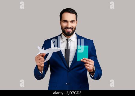 Ritratto di positivo felice uomo barbuto sorridendo e mostrando passaporto e aereo di carta a macchina fotografica, turismo, indossando tuta di stile ufficiale. Studio interno girato isolato su sfondo grigio. Foto Stock