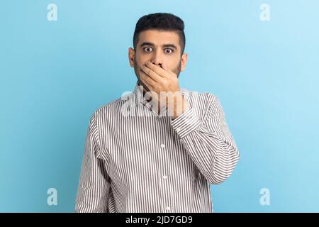 Non lo dico. Ritratto di stupito uomo d'affari che copre la bocca con la mano, mantenendo segreto, terrorizzato con notizie scioccanti, indossando camicia a righe. Studio interno girato isolato su sfondo blu. Foto Stock