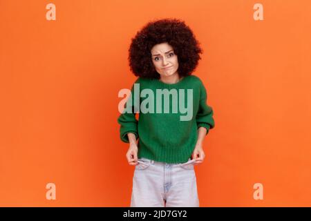 Sono in bancarotta. Donna povera infelice con acconciatura afro in maglione verde mostrando tasche vuote e guardando frustrato di prestiti e debiti, non ha soldi. Studio interno girato isolato su sfondo arancione Foto Stock