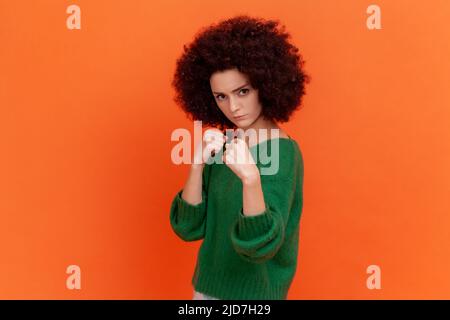 Ritratto di donna aggressiva con acconciatura Afro indossando un maglione verde casual in piedi con i pugni rialzati pronto a combattere, boxe. Studio interno girato isolato su sfondo arancione. Foto Stock