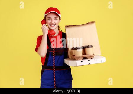 Ritratto di felice consegna donna parlare telefono fisso e tenere il caffè con pizza, chiamare per ordinare fast food, indossare tute e tappo rosso. Studio interno girato isolato su sfondo giallo. Foto Stock