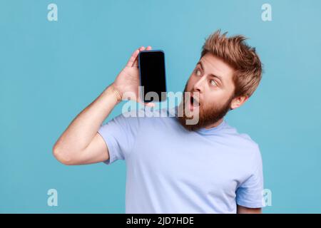 Ritratto di uomo bearded in piedi tenendo il telefono cellulare e guardando a sorpresa il gadget display con spazio vuoto, tecnologia moderna. Studio interno girato isolato su sfondo blu. Foto Stock