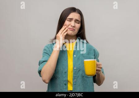 Giovane donna malata con capelli scuri in piedi e toccando la sua guancia perché sensazione di dolore sul dente, dopo aver bevuto tè caldo, indossare giacca casual stile. Studio interno girato isolato su sfondo grigio. Foto Stock