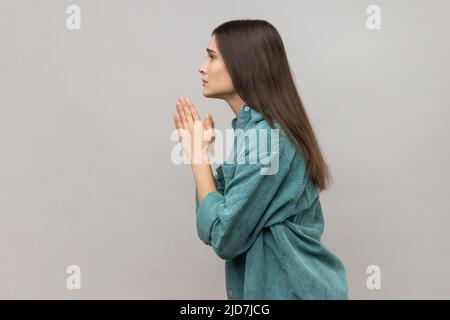 Prego, sto pregando. Vista laterale della donna che tiene le braccia nel gesto di preghiera e chiede perdono, sensazione di dispiacere per errore, indossare giacca casual stile. Studio interno girato isolato su sfondo grigio. Foto Stock