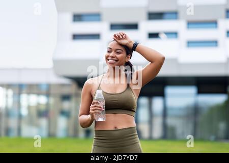 Stanca giovane donna nera in abbigliamento sportivo che fa una pausa durante il jogging, strofinando la fronte, bere acqua al parco cittadino Foto Stock