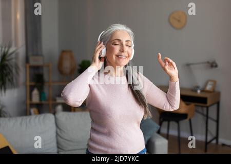 Happy Aged donna europea dai capelli grigi in cuffie wireless ascolta la musica preferita e gode di tempo libero Foto Stock