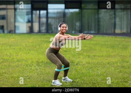 Giovane donna afroamericana sportiva che fa squat con elastico, ascoltando musica in cuffia al parco cittadino Foto Stock