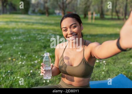 Giovane vlogger nero femmina che prende selfie o parla con i seguaci sul cellulare, facendo yoga al parco della città Foto Stock