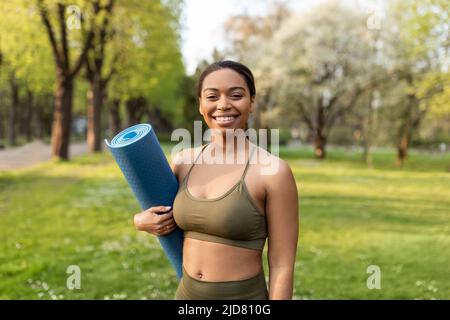 Felice giovane donna nera in abiti sportivi con tappeto fitness, sorridente alla macchina fotografica, pronta per lo yoga al parco urbano Foto Stock
