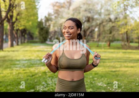 Gioiosa giovane donna nera in abiti sportivi in posa con corda di salto al parco. Concetto di stile di vita attivo Foto Stock