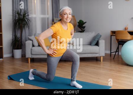 Ritratto a tutta lunghezza di donna sportiva anziana che si tiene in forma, esercitandosi sul tappetino yoga in salotto e sorridendo alla macchina fotografica Foto Stock