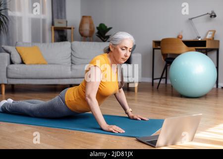 Donna anziana attiva facendo sfinge cobra posa o ascendente cane asana, excercising su tappeto e utilizzando il portatile Foto Stock