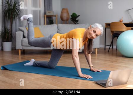Fitness domestico in linea. Donna anziana che si esercita sul video sportivo su un portatile in salotto, guardando la fotocamera e sorridendo Foto Stock
